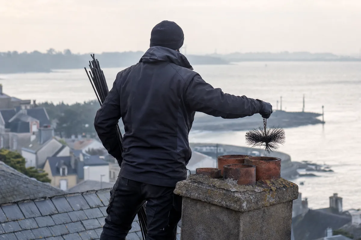 Ramoneur de dos sur un toit d&rsquo;ardoise face à l&rsquo;estuaire, cannes de ramonage à la main